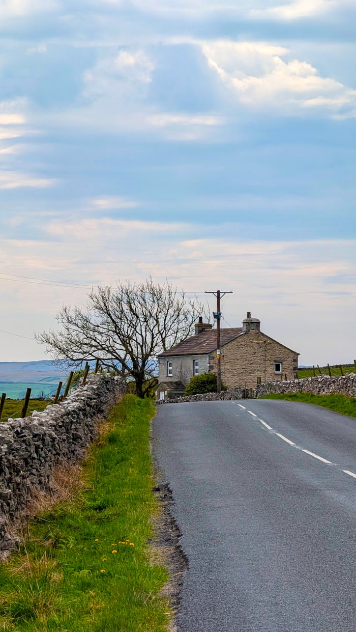 Scenic rural road with stone house and tree in the English countryside under a cloudy sky.